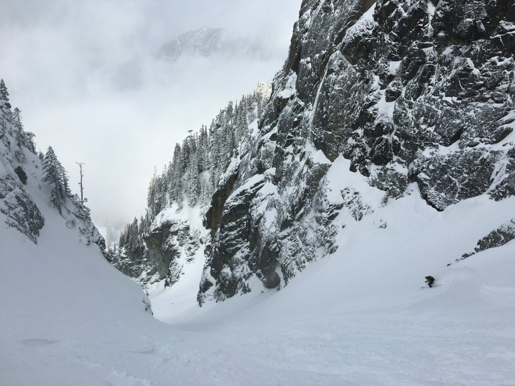 A view of a short and narrow mountain pass surrounded by granite tree-covered mountains with a backcountry skier making their way towards it.