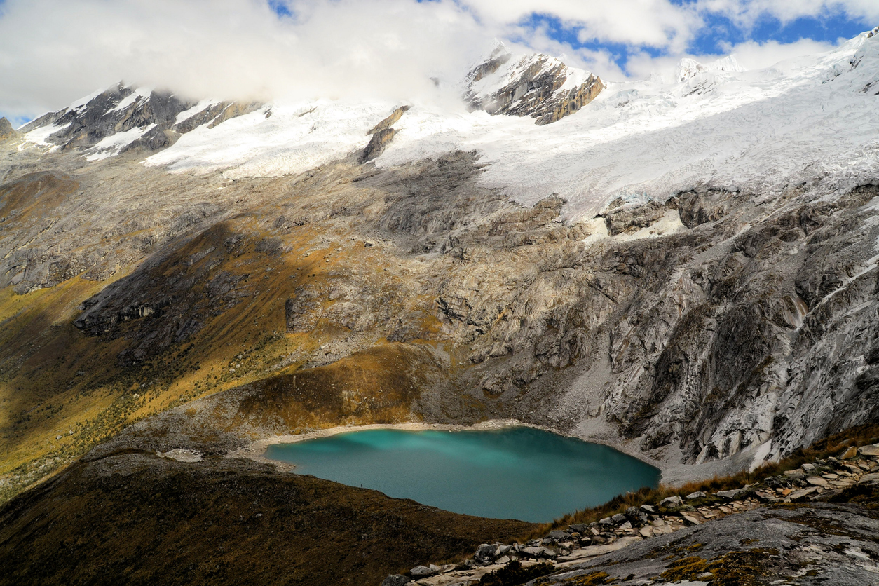 View of a turquoise lake beneath glacier-wrapped mountain peaks in Punta Union Pass. Tundra vegetation can be seen as well.