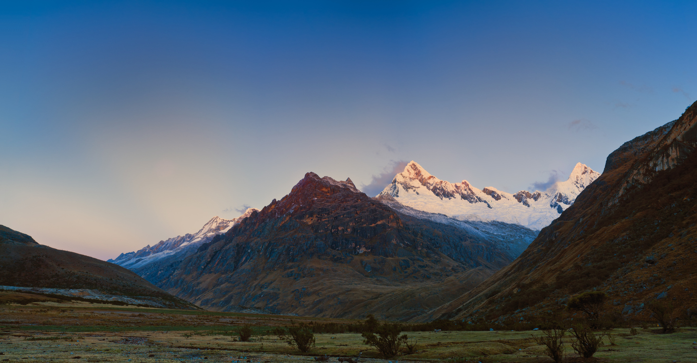 View of an Andean plateau during sunsets. It is covered in mountain vegetation and surrounded by lower hills with very high mountain peaks covered in snow in the distance.