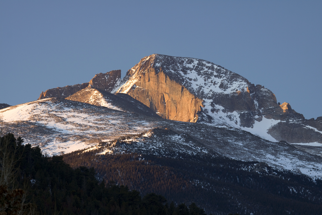 Morning sun light hits the east face or the sheer cliff known as "The Diamond" of the snow capped Longs Peak rising to 14,255 feet, in Rocky Mountain National Park, Colorado. Thick forest covers the lower elevations of the beautiful mountain.