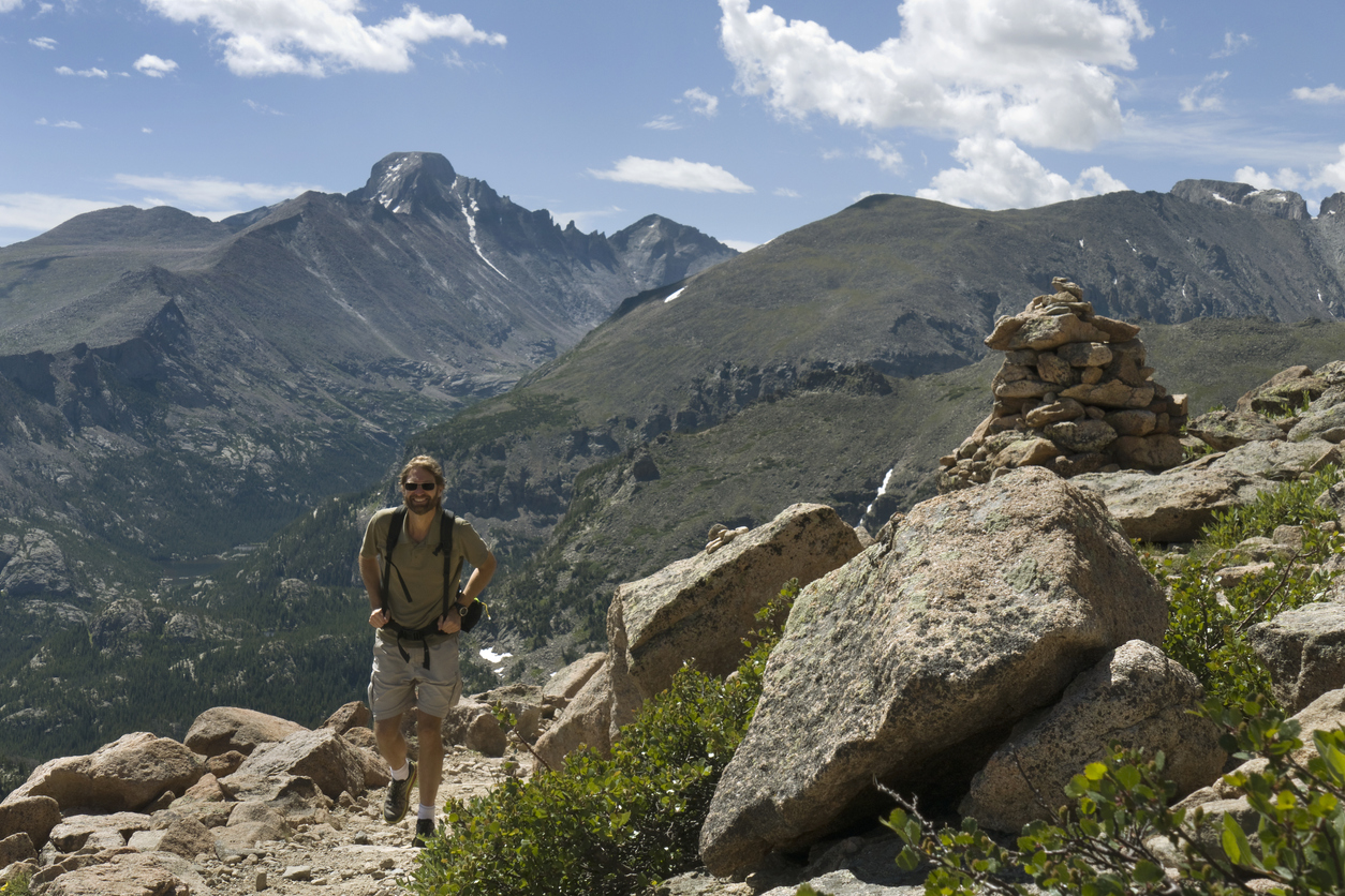 As clouds drift past the 14,255 foot Longs Peak and the rock formations known as the Keyboard of the Winds a bearded man with a backpack hikes up the Flattop mountains trail past a cairn and granite rocks in Rocky Mountain National Park, Colorado.