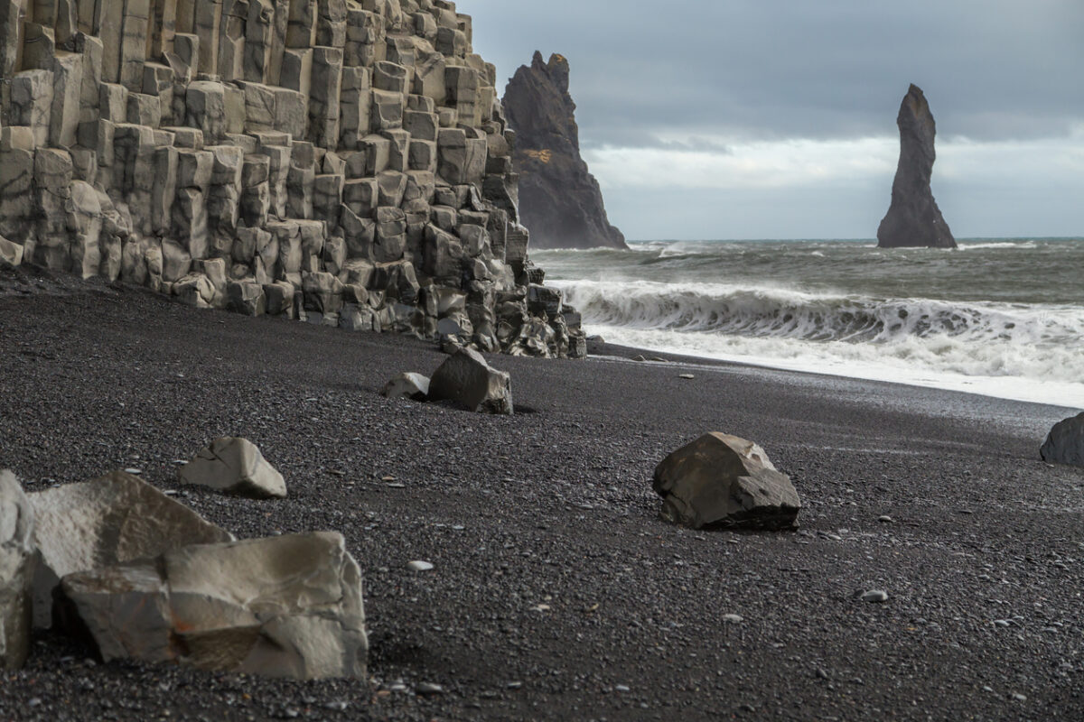 Basalt rock formations on the volcanic black sand beach at Vik in Iceland. The cliff face is columns of hexagonal rocks. Rock stacks are in the sea. Large waves and spray is being whipped up from the ocean. Rocks lie on the sand and the sky is cloudy and moody.