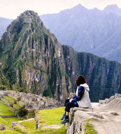 Woman sitting on a ledge and overlooking the Inca ruins of the city of Machu Picchu seen in the background.