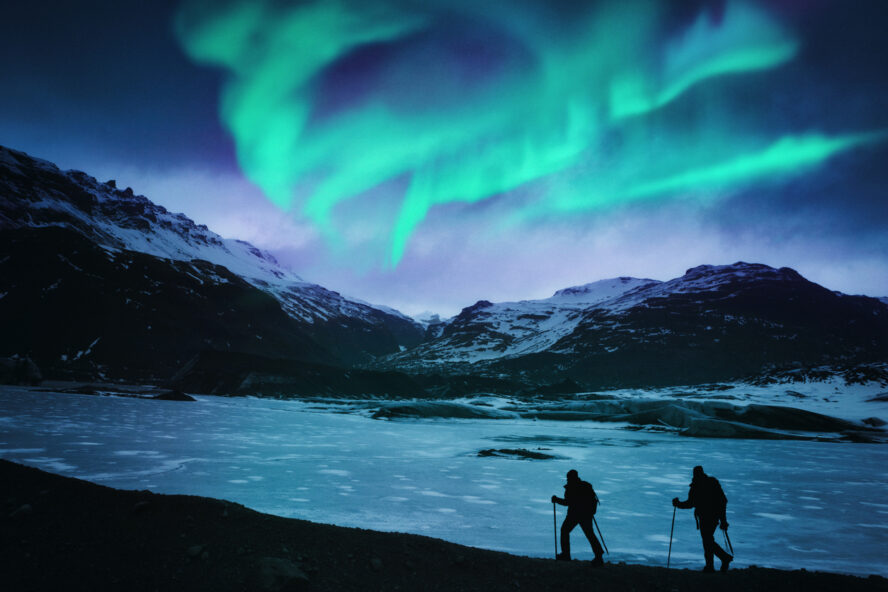 Winter hikers along a frozen lake and bathed in neon green northern lights