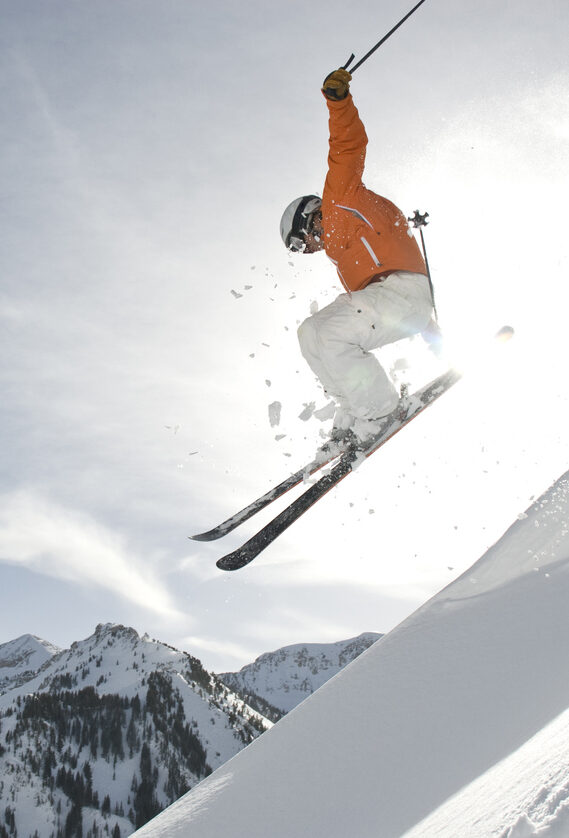 Skier jumping over a snowy ridge with sun illuminating the trailing snow dust. Shot in Utah with professional athlete wearing orange jacket and white pant, helmet and goggles.