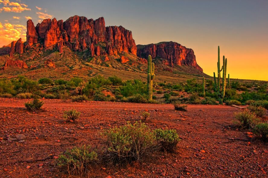 Scattered patches of desert scrub and saguaro cacti dominated by the sharp peaks of the Superstition Mountains dyed orange by the sunset.