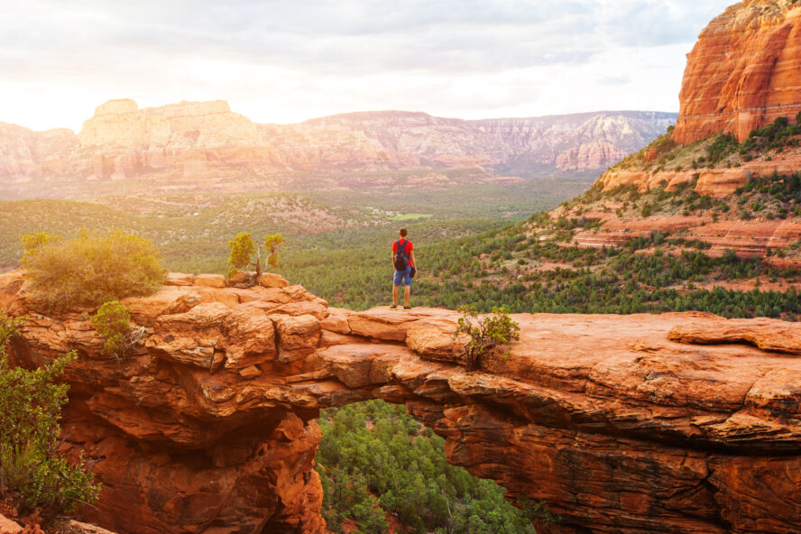 A person standing on Sedona’s Devil’s Bridge—a hanging sandstone ledge overlooking a vast forested landscape surrounded by mountains and buttes.