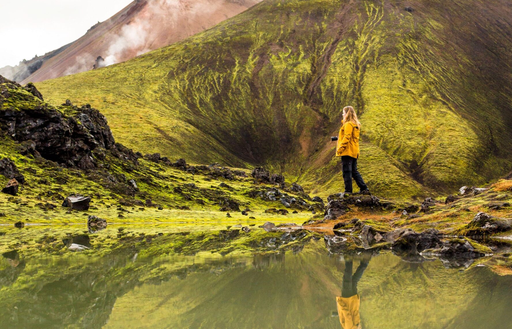 A woman standing next to a lake with mossy green mountain in the background
