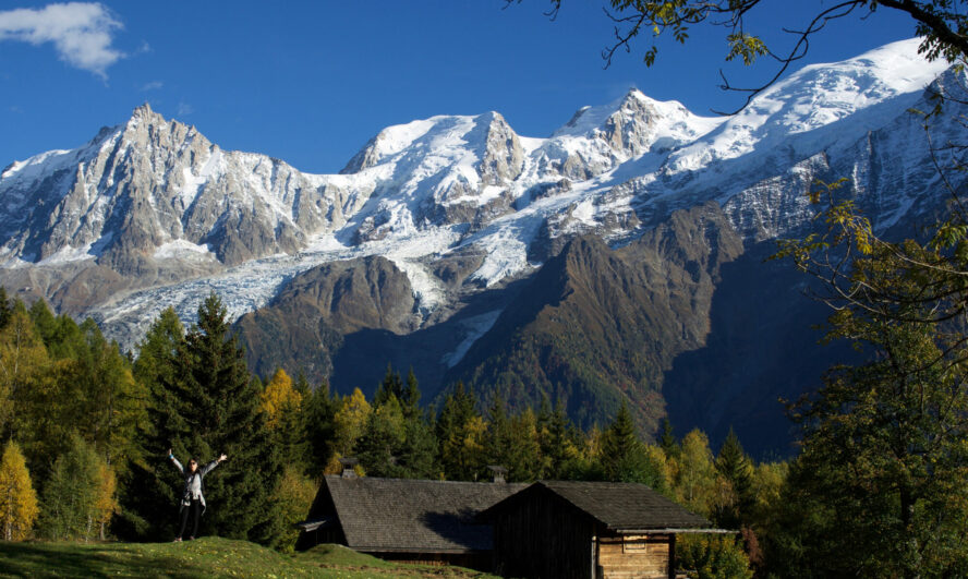A hiker on the Chamonix Renaissance Walk in Les Houches, with the Alps and a cabin in the background.