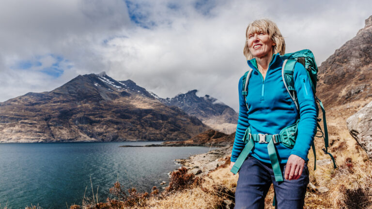A smiling hiker walking along the shore of Loch Coruisk, the Cuillin Hills in the background