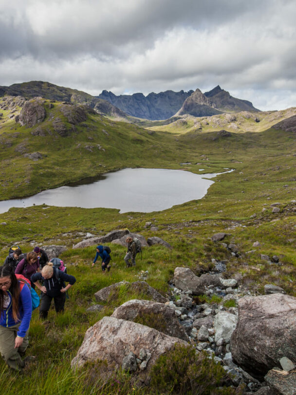 A smiling hiker walking along the shore of Loch Coruisk, the Cuillin Hills in the background