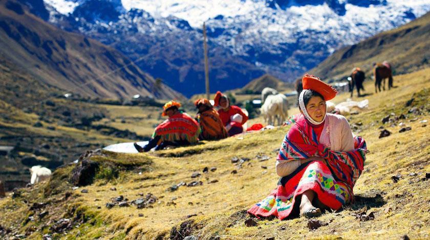 Close up of a woman sitting on grass in a traditional colourful Peruvian costume and several other people sitting behind her, looking into the distance and mountains