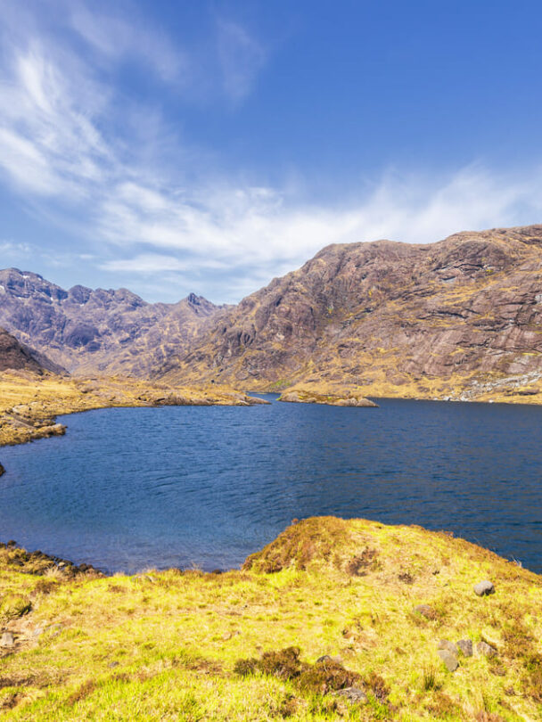 A smiling hiker walking along the shore of Loch Coruisk, the Cuillin Hills in the background