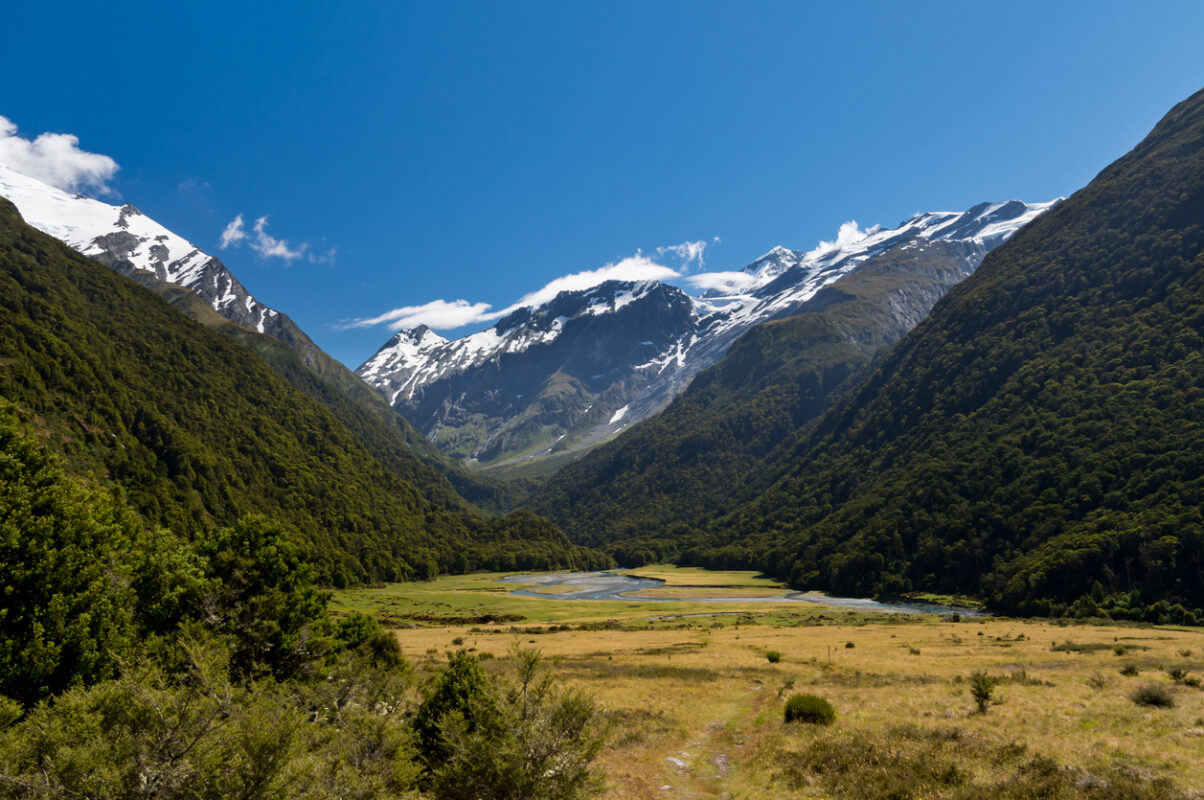 River valley surrounded by steep tree-covered mountains and one particularly high snow-covered mountain in the background. 