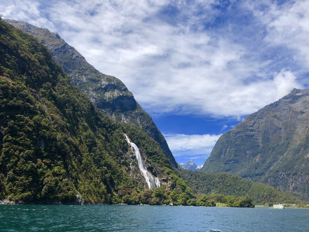 Sea-view of steep tree-covered cliffs and a waterfall with a snow-covered mountain peak in the distance.