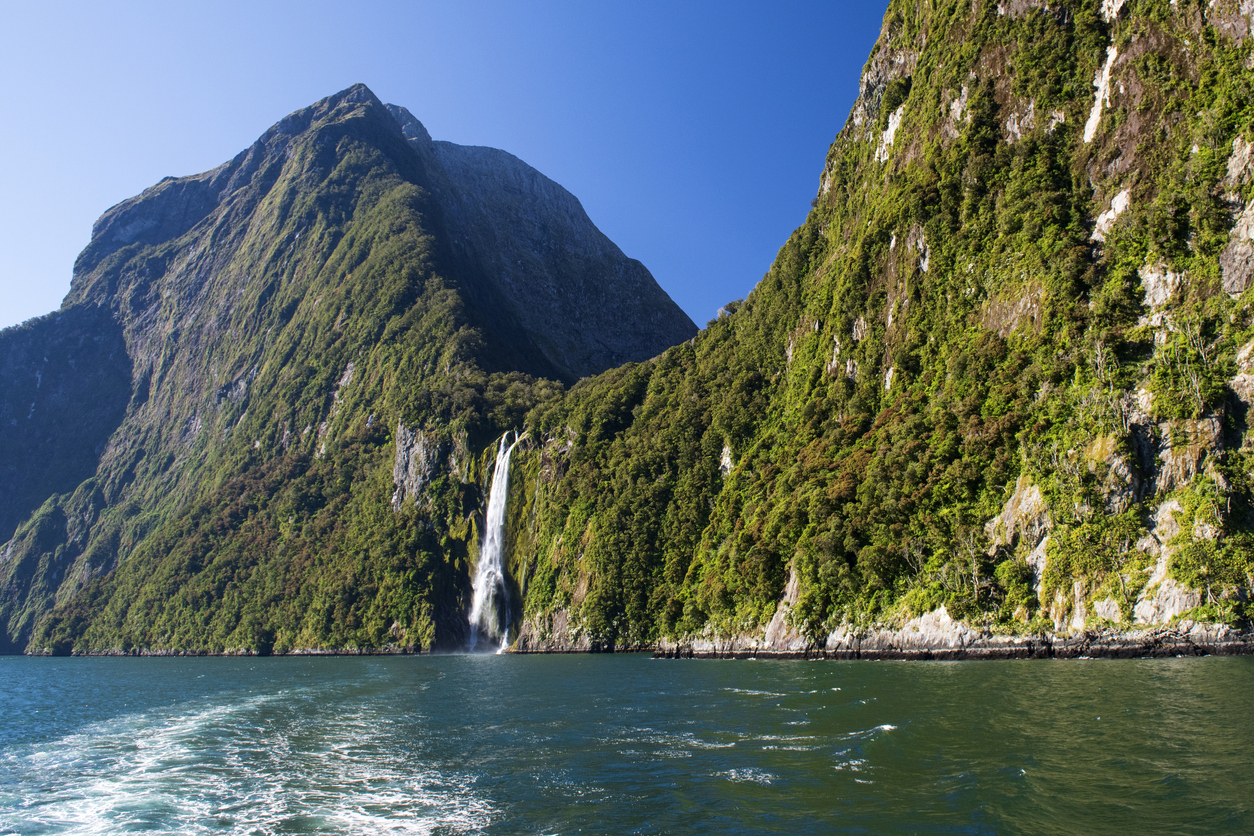 View of a tree-covered fjord wall from the water with a waterfall.