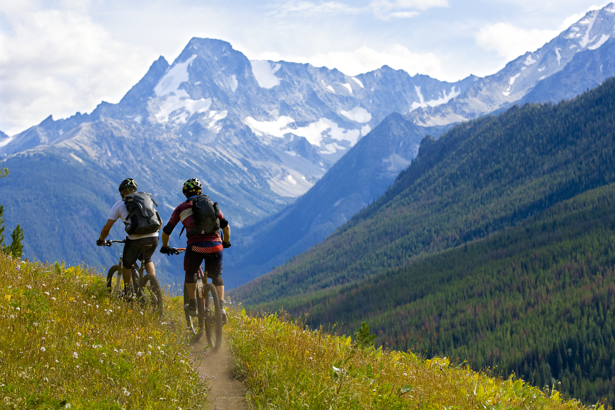 Two riders biking on a high-altitude trail with snow-covered mountain peaks and a valley with a forest in the distance.