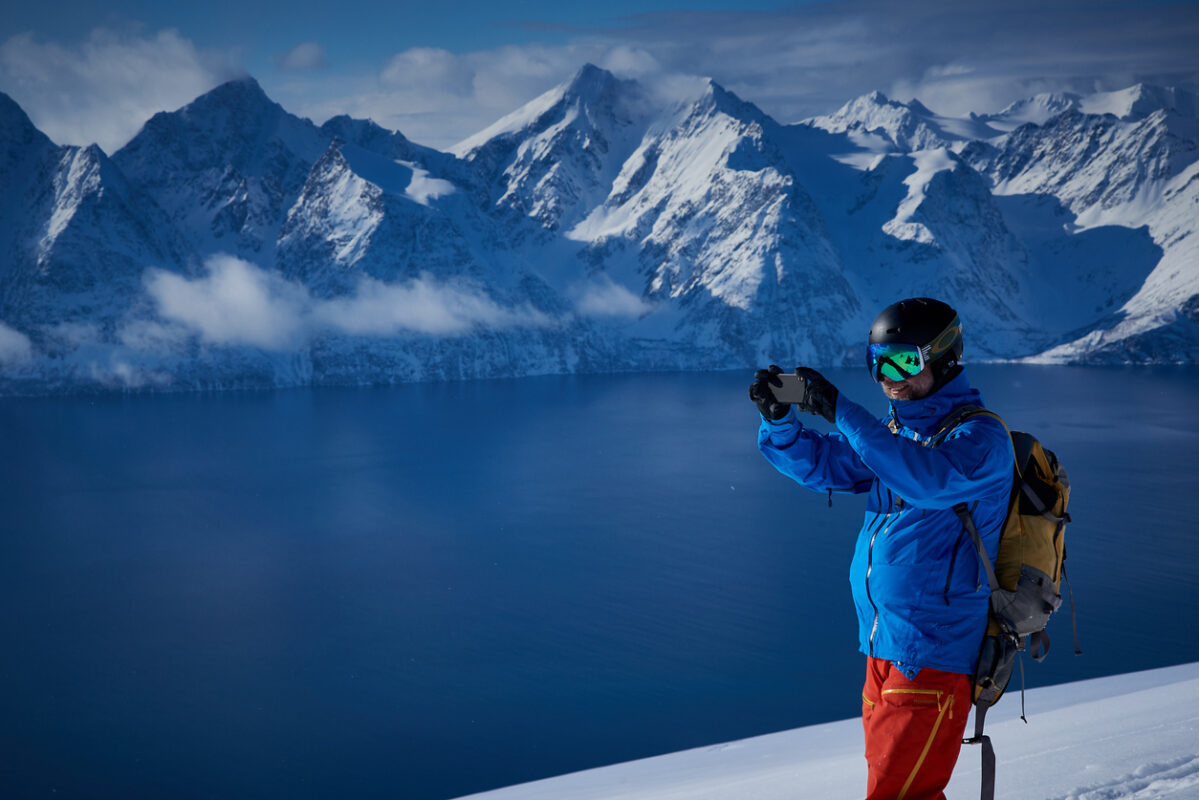 A skier on the right side of the picture is taking a photo during a skiing tour in the Lyngen Alps. Behind him there is a view of the ocean and snow-covered mountains. 