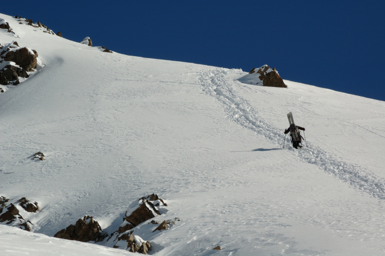 A backcountry skier is moving uphill on the slope of a volcano. The person is very small in comparison to the vast snowy landscapes