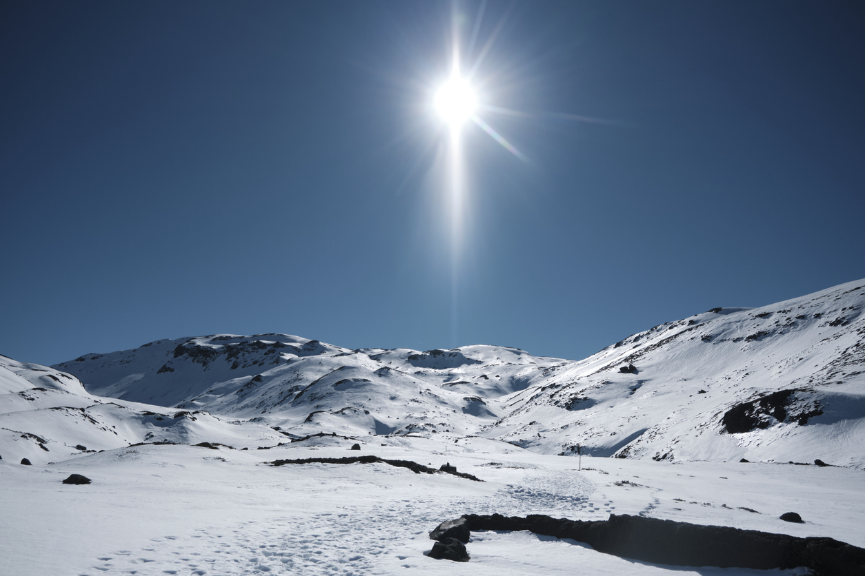 View of the mountain landscape with vast terrain covered in snow and low hills in the background. The sky is devoid of clouds and we can see the sun.
