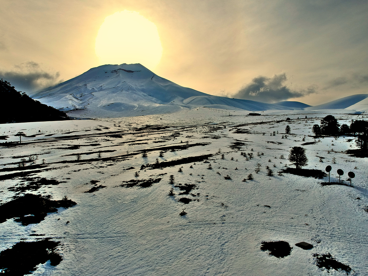 View of a snow-covered plain speckled with trees. A volcanic cone is rising abruptly from the plain in the distance. The sun is about to set behind the volcano. It is covered by thin clouds. There are warmly-colored icy reflections all over the image.