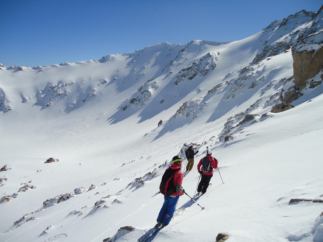 Three backcountry skiers skinning up the snow-covered rugged slope in north Patagonia. The slopes encircle a small basin.