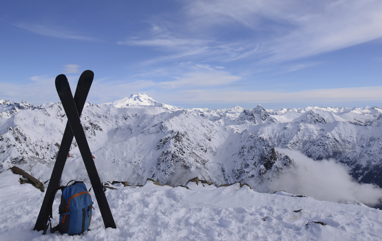 View of the snow-dusted mountains atop of a canyon with clouds. In the front of the picture there are skis and a backpack
