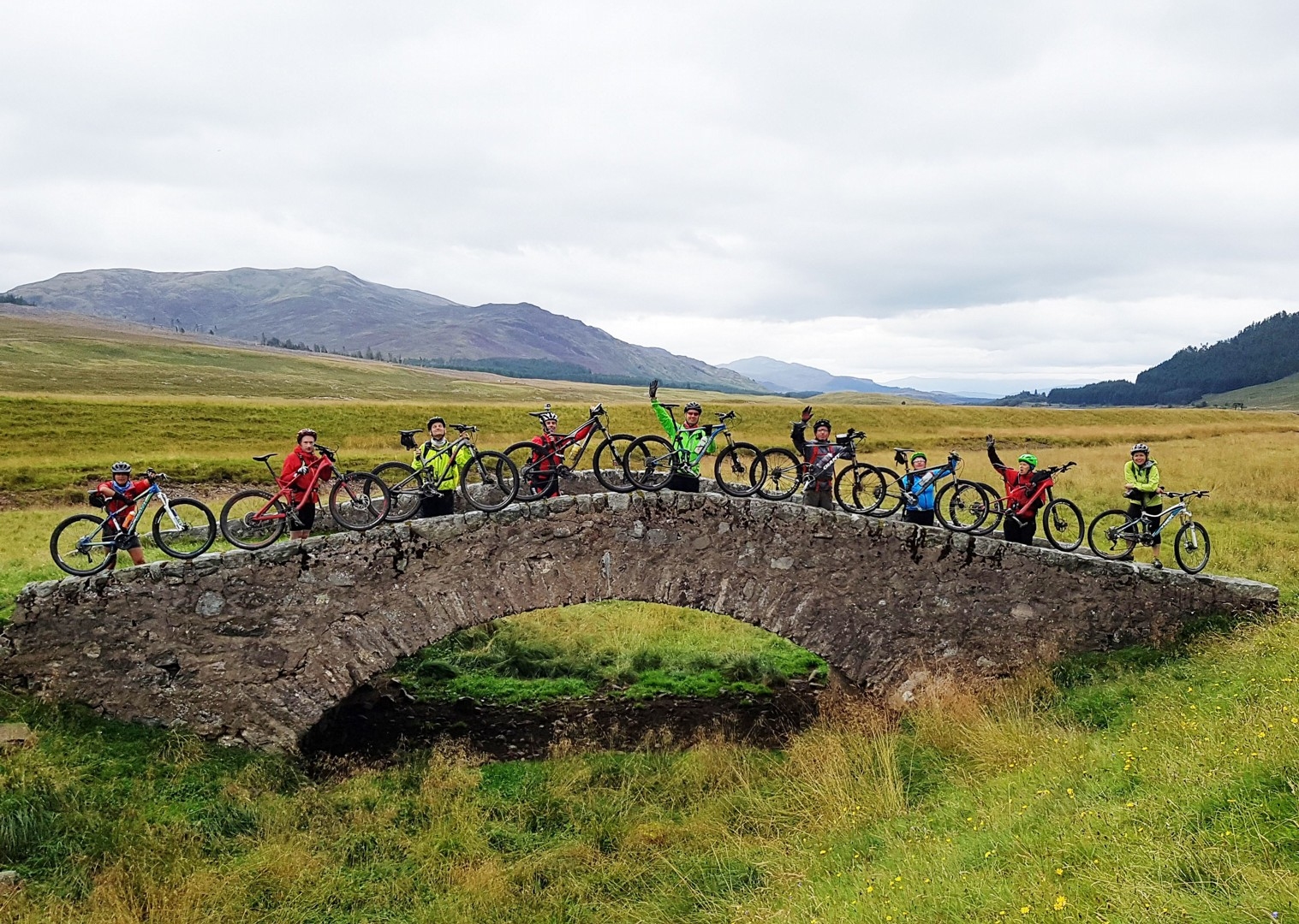 A group of mountain bikers posing for a photo on a stone bridge in the Scottish Highlands.